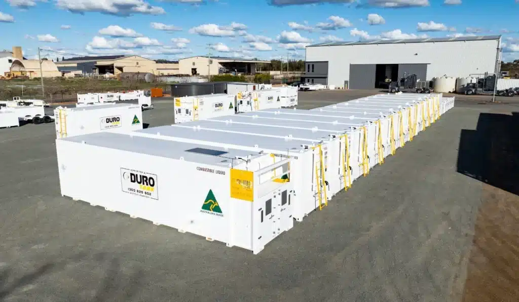 Multiple white Durotank self-bunded diesel fuel tanks lined up in a manufacturing yard, with a warehouse and trucks in the background.