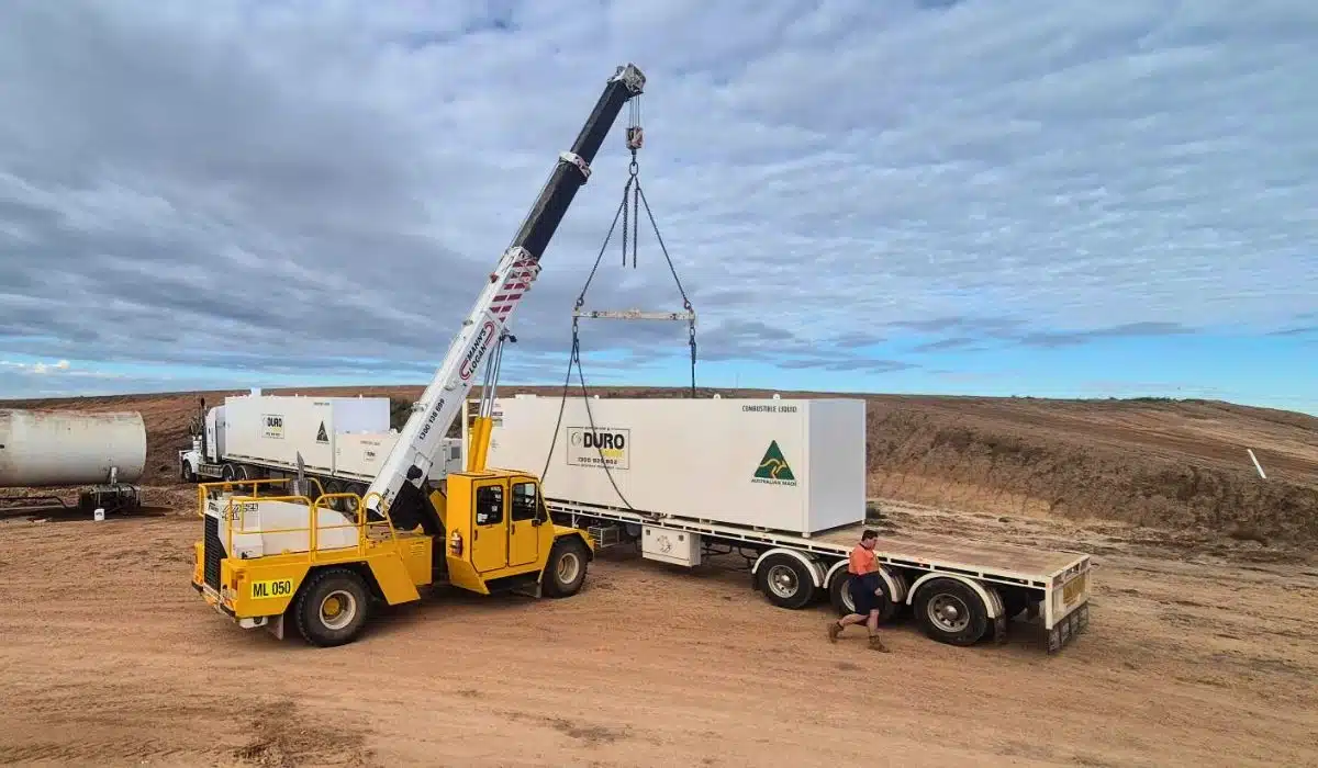 A yellow mobile crane lifting a white Durotank diesel fuel tank onto a flatbed trailer at an outback worksite, with a worker standing nearby.
